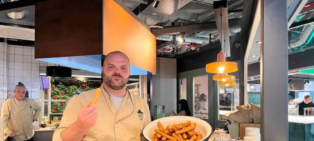 Eine Person präsentiert ein Gericht im Innenbereich der Markthalle im Königshof; eine große Schüssel mit Essen ist sichtbar.